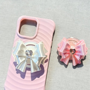 May include: Pink phone case with a white bow and a heart-shaped jewel. A matching pink bow phone grip is also shown. The phone case has a ridged design. The bows are made of satin-like material.
