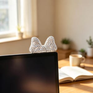 May include: A decorative bow-shaped ornament covered in clear, reflective crystals. The bow is positioned on the top edge of a black laptop screen. The crystals catch the light, creating a sparkling effect across the surface of the bow accessory.