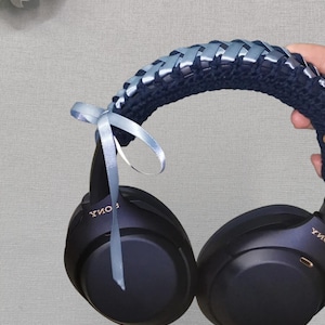 May include: Black over-ear headphones with a navy blue crocheted headband. The headband is decorated with light blue ribbon woven through the crochet. A light blue ribbon bow is tied to the headphones.