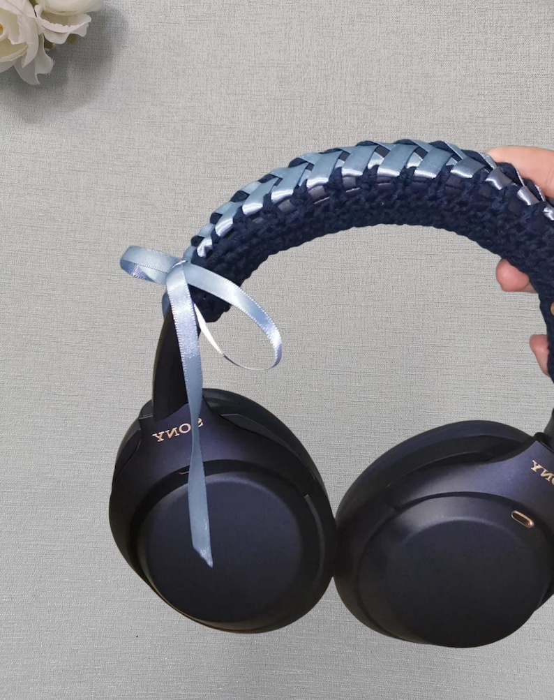 May include: Black over-ear headphones with a navy blue crocheted headband. The headband is decorated with light blue ribbon woven through the crochet. A light blue ribbon bow is tied to the headphones.