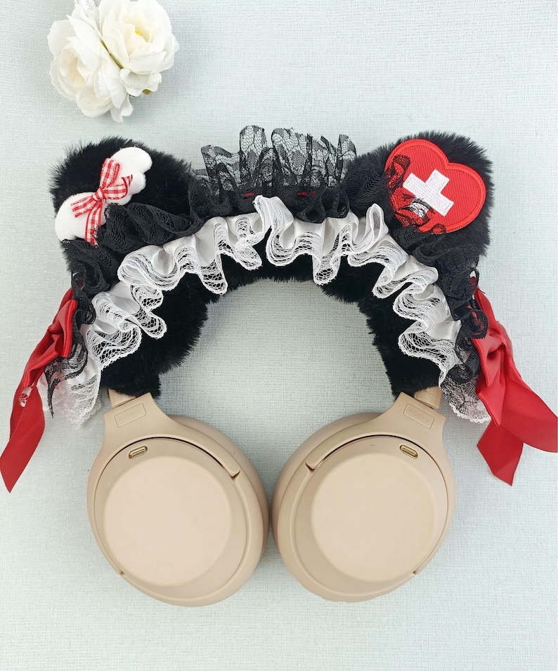May include: Black faux fur headphone accessory with red and white accents. Features a red heart with a white cross, red bows, and layers of black lace and white ruffled trim. The headphones are beige.