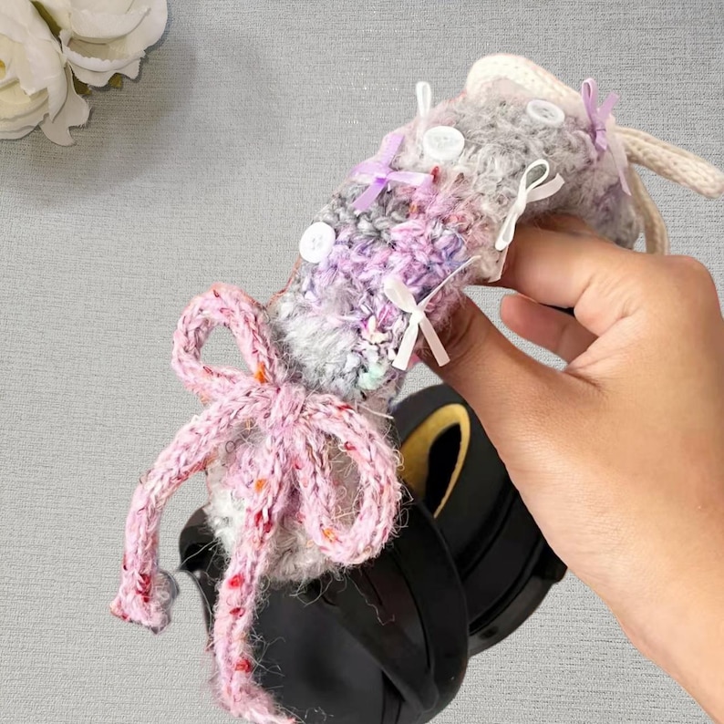 May include: A handmade, multi-colored crochet item with a large pink bow and small white bows. The item has a mix of gray, purple, and white yarn, with white buttons and a cream-colored cord. The item is held in a hand.