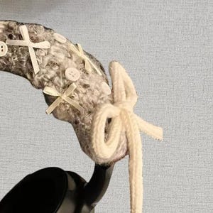 May include: Headphones with a gray and white knitted headband, adorned with white bows and buttons. A cream-colored, knotted cord hangs from the headband. The headphones are black.