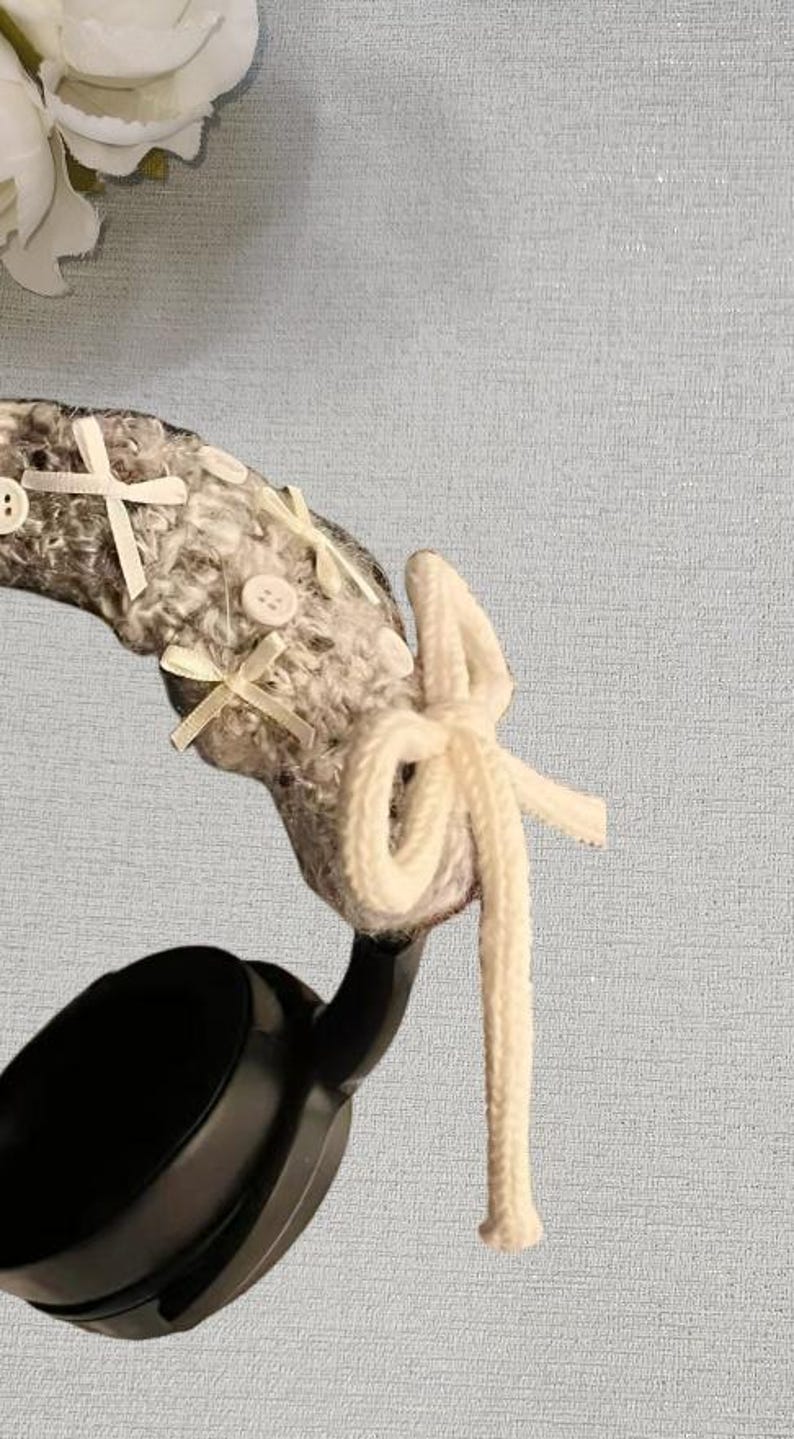 May include: Headphones with a gray and white knitted headband, adorned with white bows and buttons. A cream-colored, knotted cord hangs from the headband. The headphones are black.