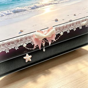 May include: A black electronic device decorated with a pink and white fabric bow, white star-patterned lace trim, and a dangling pearl-like charm. A small, light-coloured star-shaped embellishment is attached to the front of the device.