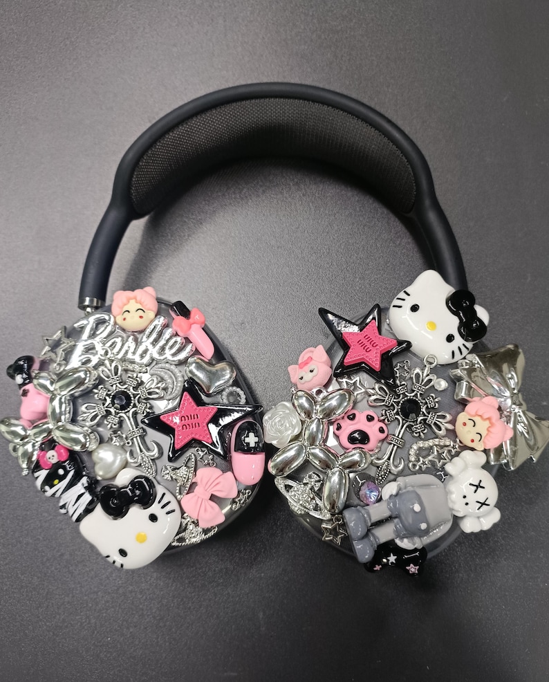 K&ouml;nnte beinhalten: Schwarzer Kopfh&ouml;rer, verziert mit verschiedenen Charms, darunter Hello Kitty, Sterne, Schleifen und das Wort "Barbie". Die Charms sind in Rosa, Silber und Wei&szlig; gehalten und schaffen ein verspieltes und dekoratives Design.