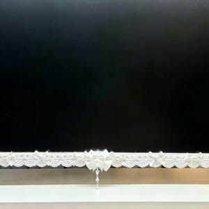 以下が含まれることがあります： 複雑なレース模様、小さなリボン、両端の翼のようなアクセントが特徴の白い装飾フレーム。スクリーンやモニター用のこの水平なディスプレイピースの中央のリボンには、小さなクリスタル風のチャームがぶら下がっています。