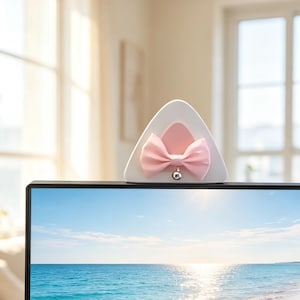 Puede incluir: Una cubierta de cámara web triangular blanca con una forma de oreja interna rosa, un lazo de tela rosa unido al centro y una pequeña campana plateada que cuelga de la parte inferior del lazo, montada en el borde superior de un monitor de PC.