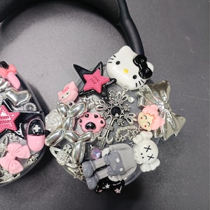 K&ouml;nnte beinhalten: Schwarzer Kopfh&ouml;rer, verziert mit verschiedenen Charms, darunter eine Hello Kitty Figur, ein rosa Stern, eine graue Figur und silberne Verzierungen. Die Charms sind um die Ohrmuschel angeordnet und schaffen ein einzigartiges Accessoire.