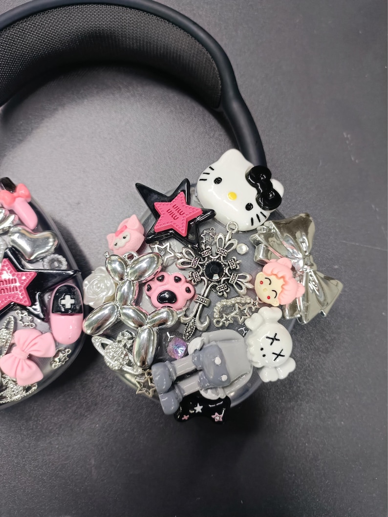 K&ouml;nnte beinhalten: Schwarzer Kopfh&ouml;rer, verziert mit verschiedenen Charms, darunter eine Hello Kitty Figur, ein rosa Stern, eine graue Figur und silberne Verzierungen. Die Charms sind um die Ohrmuschel angeordnet und schaffen ein einzigartiges Accessoire.