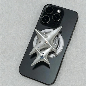 Pode incluir: Um smartphone preto com um suporte de telefone prateado em forma de estrela anexado. A pega do telefone tem um acabamento metálico e é adornada com pequenos enfeites em forma de estrela coloridos. As lentes da câmara do telefone são visíveis.