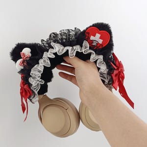 May include: Black faux fur cat ear headphones with red bows and a red heart-shaped patch with a white cross. The headphones have a black lace and white ruffled trim.