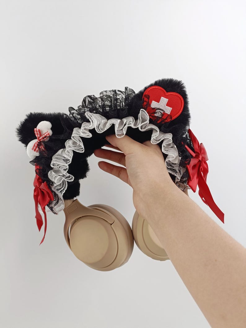May include: Black faux fur cat ear headphones with red bows and a red heart-shaped patch with a white cross. The headphones have a black lace and white ruffled trim.