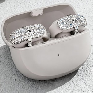 Op de afbeelding: Draadloze oortelefoons in een lichtgrijze oplaadcase. De oortelefoons zijn versierd met sprankelende strass-steentjes. De case is open en toont de oortelefoons. De achtergrond is een gestructureerd, lichtgrijs oppervlak.
