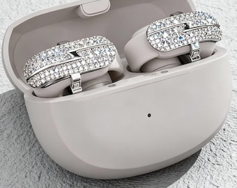 May include: A pair of wireless earbuds in a light gray charging case. The earbuds are adorned with sparkling rhinestones. The case is open, revealing the earbuds inside. The background is a textured, light gray surface.