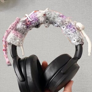 May include: Black over-ear headphones with a crocheted headband cover. The cover is a mix of pink, purple, and gray yarn, adorned with small white buttons and accents. The headphones are held by a hand.