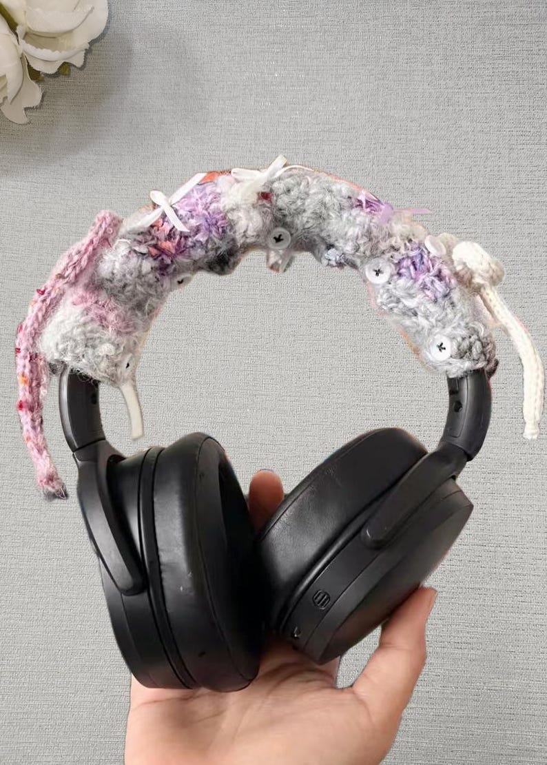 May include: Black over-ear headphones with a crocheted headband cover. The cover is a mix of pink, purple, and gray yarn, adorned with small white buttons and accents. The headphones are held by a hand.