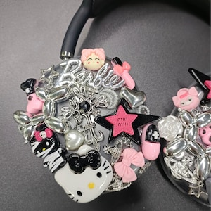 K&ouml;nnte beinhalten: Schwarzer Kopfh&ouml;rer, verziert mit dekorativen Charms, darunter ein Hello Kitty-Design, rosa Schleifen und das Wort "Barbie" in silberner Schrift. Das Design beinhaltet Sterne, Herzen und andere Verzierungen.