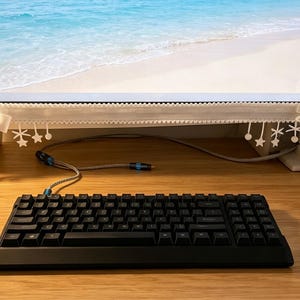 May include: A white lace monitor cover featuring decorative bows and hanging star-shaped ornaments attached to the bottom edge. The trim has a scalloped design and is positioned along the lower frame of a computer screen displaying a beach scene.