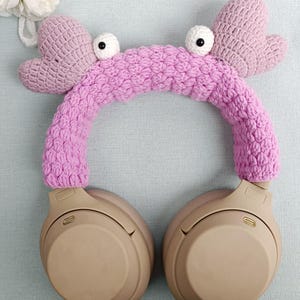 Puede incluir: Diadema de auriculares de color rosa hecha a crochet en forma de cangrejo con ojos negros. La diadema est&aacute; en un par de auriculares beige de tama&ntilde;o grande.