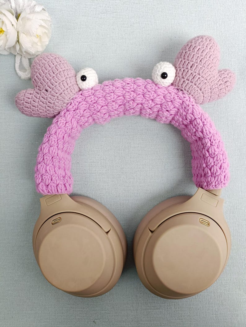 Puede incluir: Diadema de auriculares de color rosa hecha a crochet en forma de cangrejo con ojos negros. La diadema est&aacute; en un par de auriculares beige de tama&ntilde;o grande.