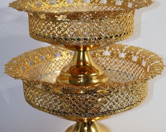 May include: A two-tiered gold-colored metal serving stand with a decorative, intricate design. The stand has a round top and a wide base.