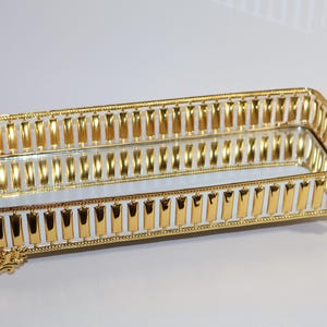 May include: A gold-colored rectangular tray with a mirrored surface and decorative detailing. The tray has a raised edge and four feet.
