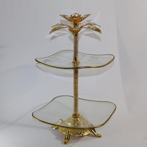 Könnte beinhalten: Ein zweistöckiger Servierständer aus Gold mit klaren Glasplatten. Der Ständer ist mit einem dekorativen goldenen Palmenbaum verziert.
