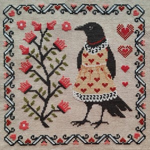 May include: Cross-stitch design featuring a black crow wearing a tan apron adorned with red hearts. A flowering tree and decorative border with hearts and floral elements surround the crow. The color palette includes red, black, tan, and green.