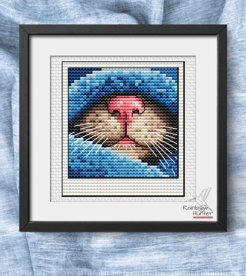 May include: Framed cross-stitch art featuring a close-up of a cat's face. The cat has a pink nose, tan muzzle, and blue fur. The artwork is in a black frame and includes the text "Rainbow Hunter".