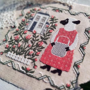 May include: A cross-stitch design depicting a sheep character in a red dress carrying a basket, next to a house and surrounded by flowers. The design includes the word "BELLE" and is framed with a decorative border.