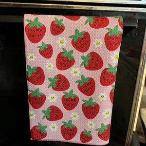 May include: A pink kitchen towel with a pattern of red strawberries, green leaves, and white daisies. Each strawberry has the text "TRUMP SUCKS" printed on it. The towel is hanging on a black oven handle.