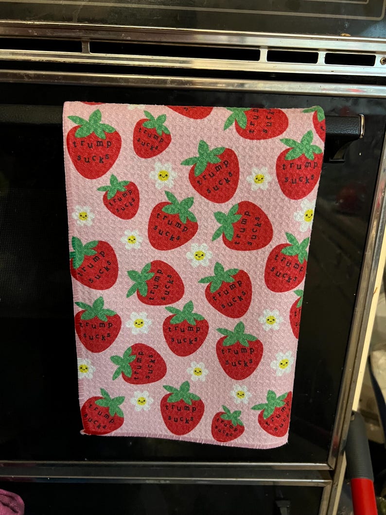 May include: A pink kitchen towel with a pattern of red strawberries, green leaves, and white daisies. Each strawberry has the text "TRUMP SUCKS" printed on it. The towel is hanging on a black oven handle.