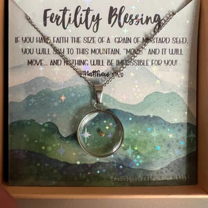 May include: A silver necklace with a clear glass pendant containing a small gold seed. The necklace is in a box with a card that reads "Fertility Blessing" and a quote from Matthew 17:20.