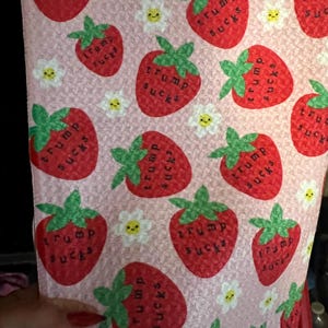 May include: Pink fabric with a repeating pattern of red strawberries with green stems and the text "trump sucks" in black. Small white flowers with yellow centers are also scattered across the fabric.