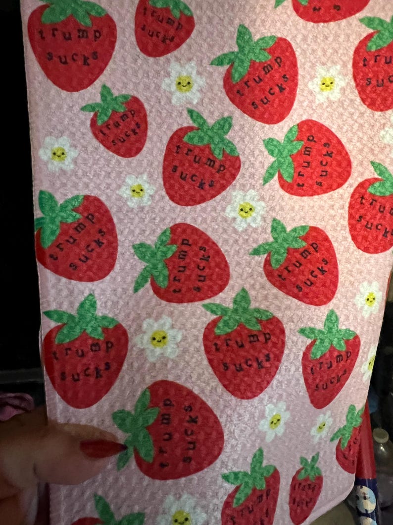 May include: Pink fabric with a repeating pattern of red strawberries with green stems and the text "trump sucks" in black. Small white flowers with yellow centers are also scattered across the fabric.
