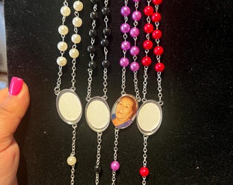 May include: Four rosaries with different colored beads: white, black, purple, and red. Each rosary has a silver cross and an oval frame. One rosary has a photo inside the frame. The rosaries are displayed on a black surface.
