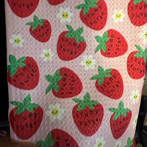 May include: Pink fabric with a pattern of red strawberries, green leaves, and white daisies. Each strawberry has the words "TRUMP SUCKS" printed on it. The daisies have yellow centers.