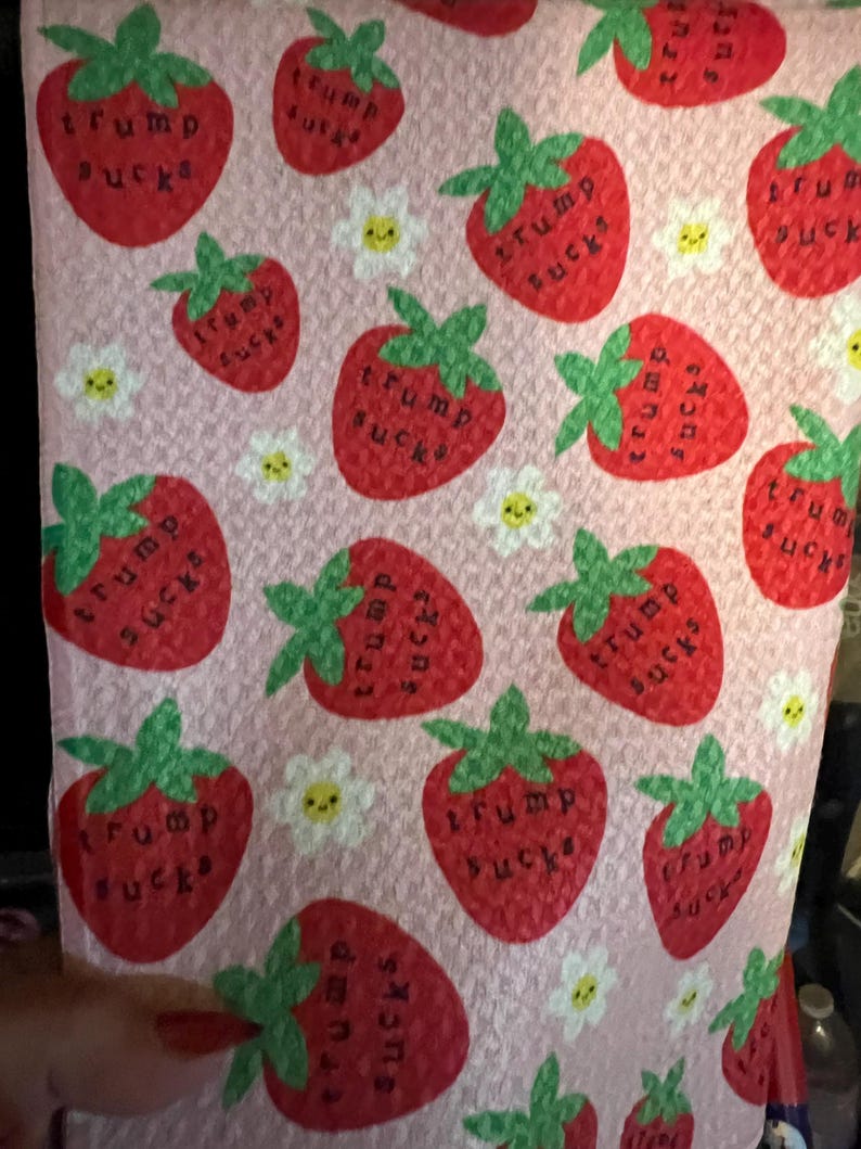 May include: Pink fabric with a pattern of red strawberries, green leaves, and white daisies. Each strawberry has the words "TRUMP SUCKS" printed on it. The daisies have yellow centers.