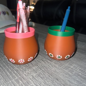 May include: Two terracotta-colored pen holders with a floral design. One has a pink rim and the other has a green rim. Pens and a pen are inside the holders. The holders are on a gray surface.