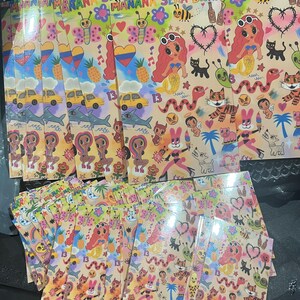 May include: A stack of colorful stickers with a variety of designs, including a mermaid, a tiger, a snake, a butterfly, and a pineapple. The stickers are printed on white paper and have a glossy finish. The text "MANANA SEKA DANI" is printed on the stickers.