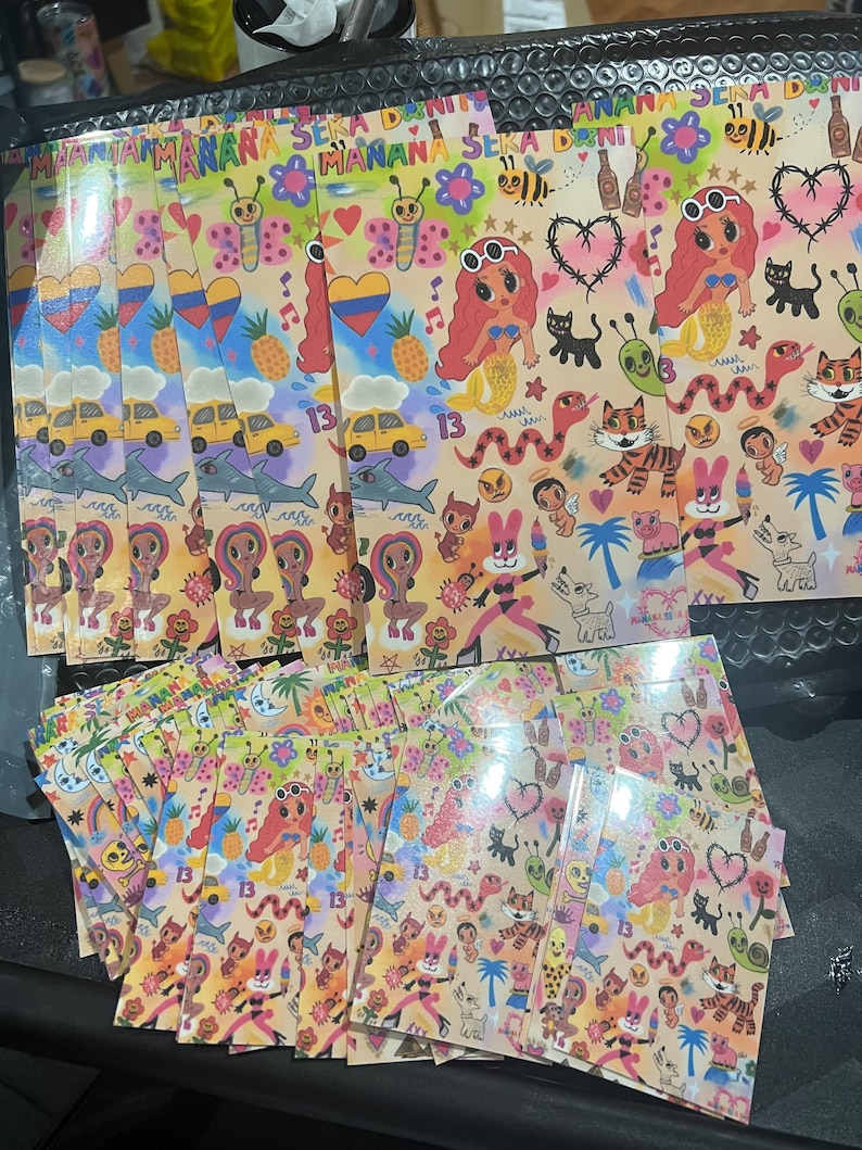 May include: A stack of colorful stickers with a variety of designs, including a mermaid, a tiger, a snake, a butterfly, and a pineapple. The stickers are printed on white paper and have a glossy finish. The text "MANANA SEKA DANI" is printed on the stickers.