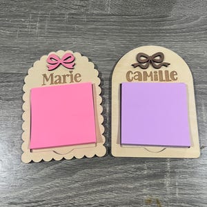 May include: Two wooden memo holders with personalized names. One has a pink bow and the name "Marie" above a pink sticky note. The other has a brown bow and the name "Camille" above a purple sticky note. Both are on a wooden surface.