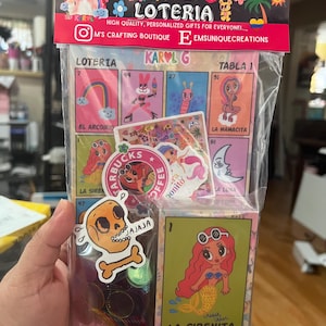 May include: A set of colorful Loteria cards featuring a mermaid, a skull, and a rainbow. The cards are packaged in a clear plastic bag with stickers and other accessories. The bag has a label that says "Karol G Loteria" and "High Quality, Personalized Gifts for Everyone!"