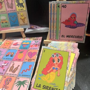 May include: A set of colorful illustrated cards with Spanish text. The cards feature various characters and images, including a mermaid, a heart, a snake, and a pineapple. The cards are numbered and have a title at the bottom. The cards are arranged in a grid pattern on a wooden easel.