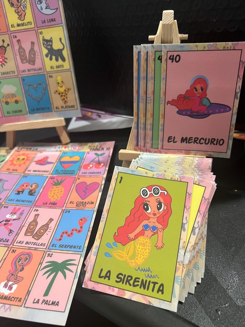 May include: A set of colorful illustrated cards with Spanish text. The cards feature various characters and images, including a mermaid, a heart, a snake, and a pineapple. The cards are numbered and have a title at the bottom. The cards are arranged in a grid pattern on a wooden easel.