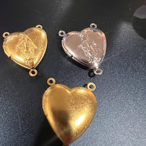 May include: Three heart-shaped lockets in gold and silver tones. Each locket features a religious figure embossed on the front and small loops for attaching a chain. The lockets are displayed on a dark surface.