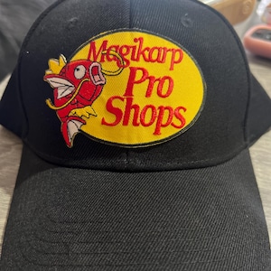 May include: Black baseball cap with a curved brim. The cap features an embroidered patch with the text "Magikarp Pro Shops" in red and yellow, along with a red cartoon fish design. The cap is made of a textured fabric.
