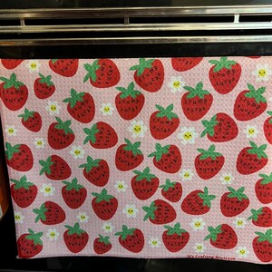 May include: Pink kitchen towel with a pattern of red strawberries, each with the text "Trump Sucks". Small white daisies are interspersed throughout the design. The towel is displayed on an oven.