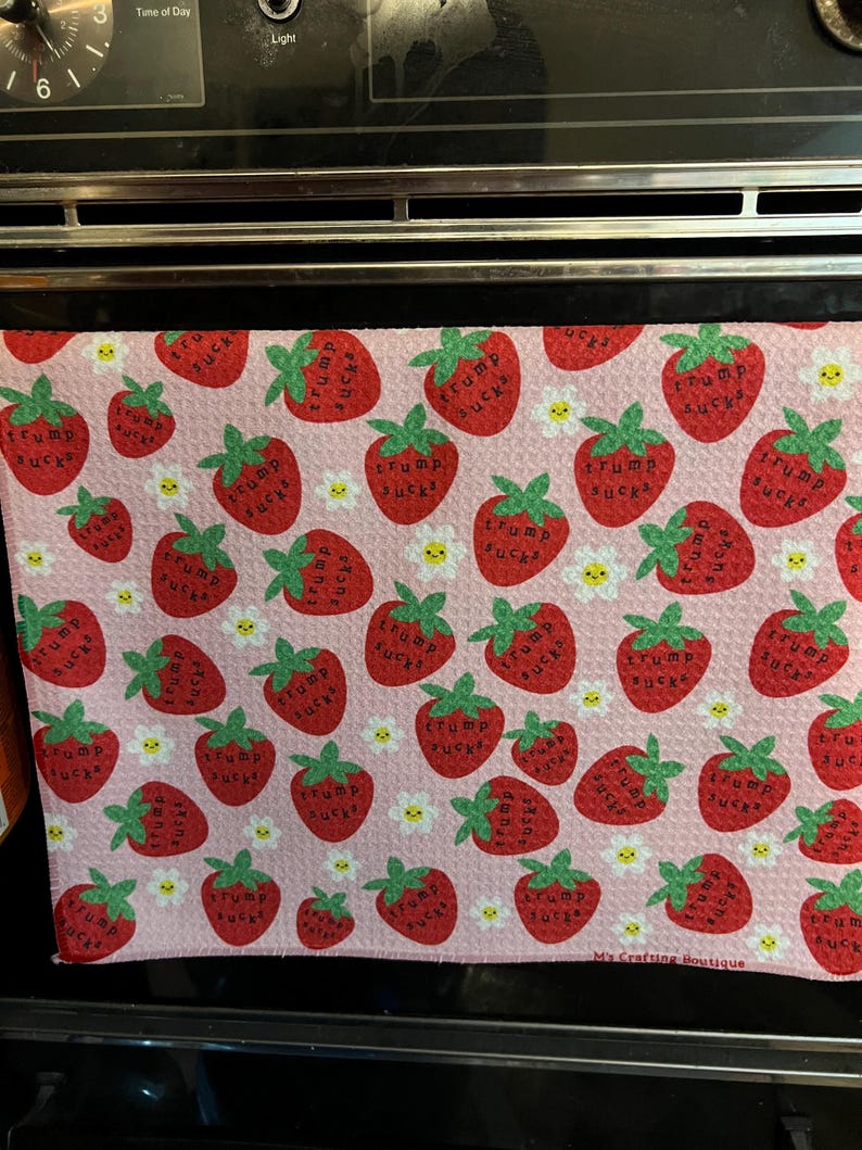 May include: Pink kitchen towel with a pattern of red strawberries, each with the text "Trump Sucks". Small white daisies are interspersed throughout the design. The towel is displayed on an oven.